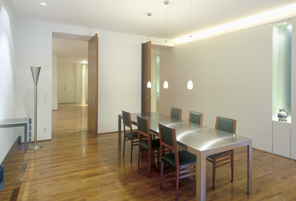 Nowoczesna jadalnia z parkietem, metalowym stołem i minimalistycznymi lampami wiszącymi, widok na otwarte drzwi do kolejnego pomieszczenia.